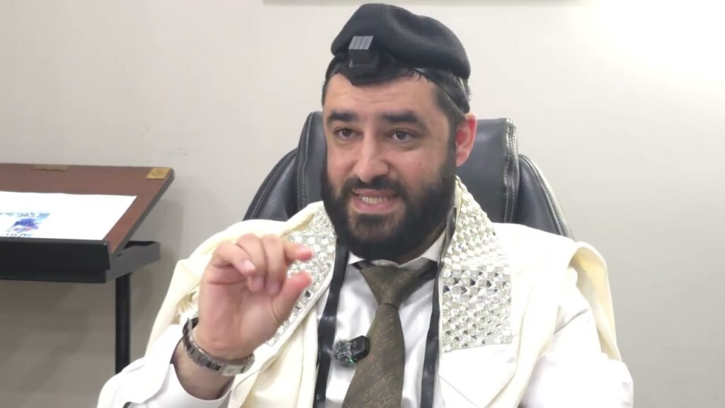 Bearded man in traditional robes and a patterned scarf sits in an office chair, speaking and gesturing with a microphone clipped to his chest.