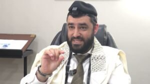 Bearded man in traditional robes and a patterned scarf sits in an office chair, speaking and gesturing with a microphone clipped to his chest.