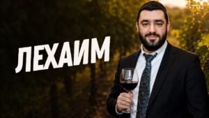Professional man in a suit holding a glass of red wine in a vineyard at sunset; large Hebrew word L'CHAIM across the left, with a small 'בס"ד' in the top-right corner.