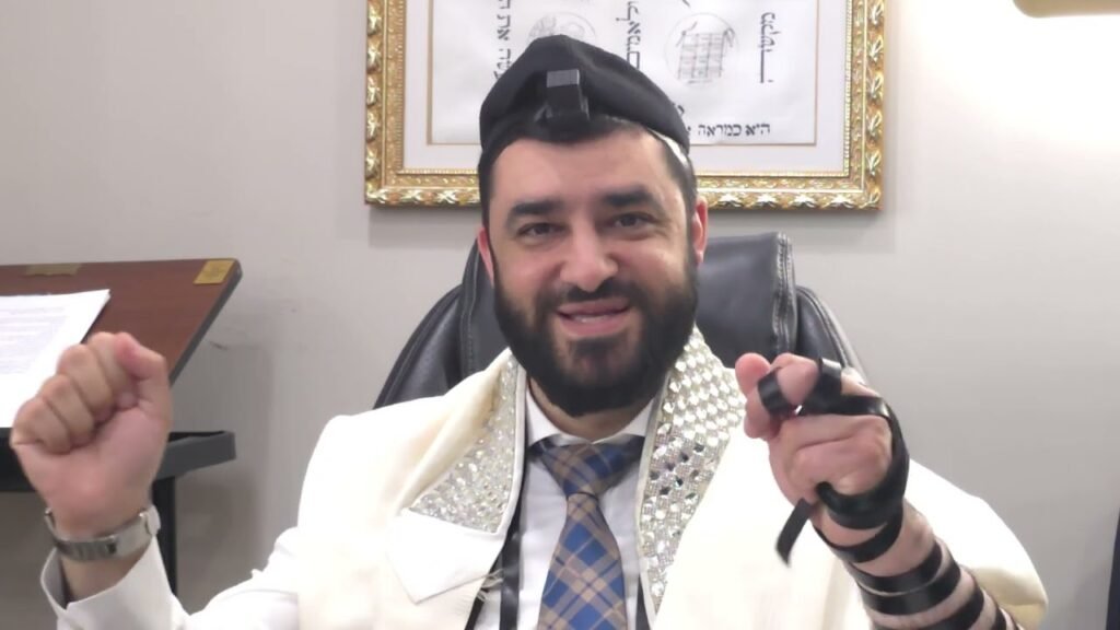 Bearded man in a white ceremonial robe with silver trim and a blue plaid tie, wearing tefillin on his head and arm, smiling at the camera.