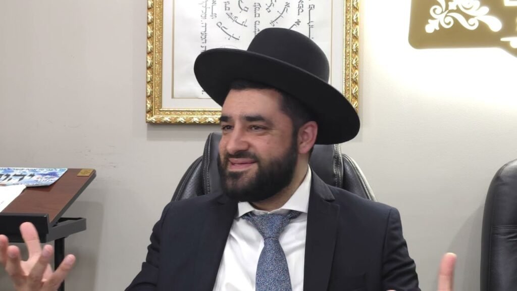 Bearded man in a dark suit and blue tie sits in an office chair, wearing a wide-brimmed black hat and gesturing with his hands.