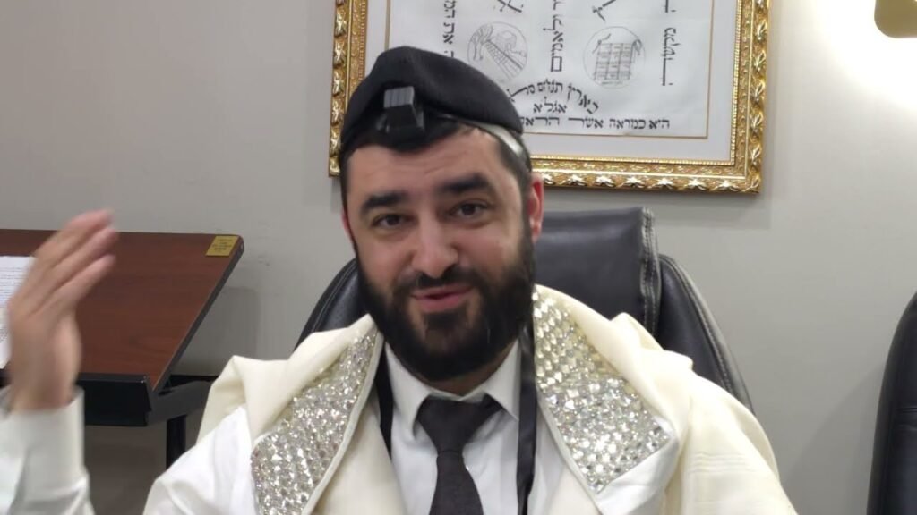Bearded man in a white prayer shawl with silver-edged trim, wearing a tefillin box on his head, speaking in an office setting.