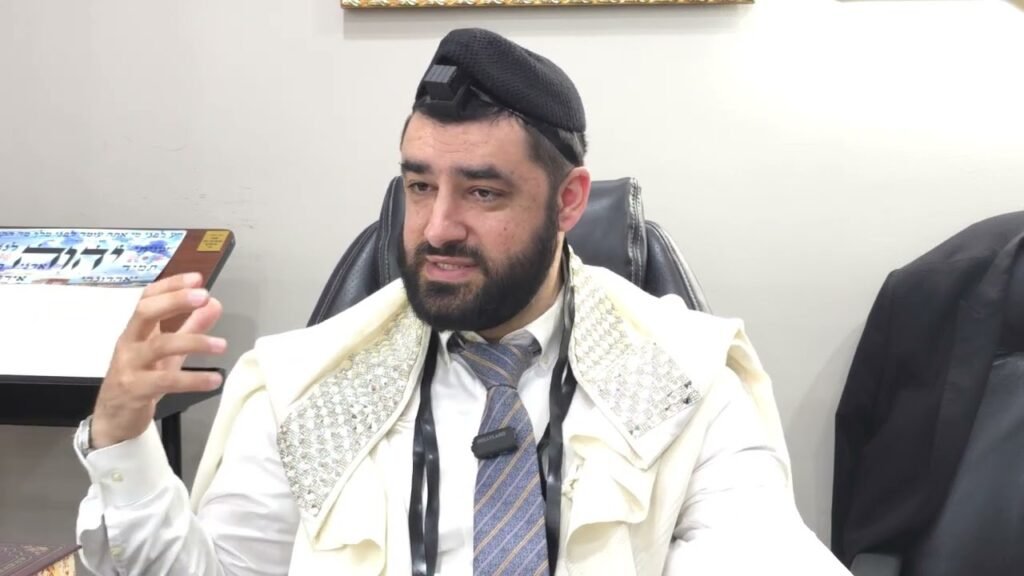 Bearded man in traditional Jewish prayer attire sits in an office chair, speaking with a hand gesture; tefillin on his head and a shawl draped over his shoulders.