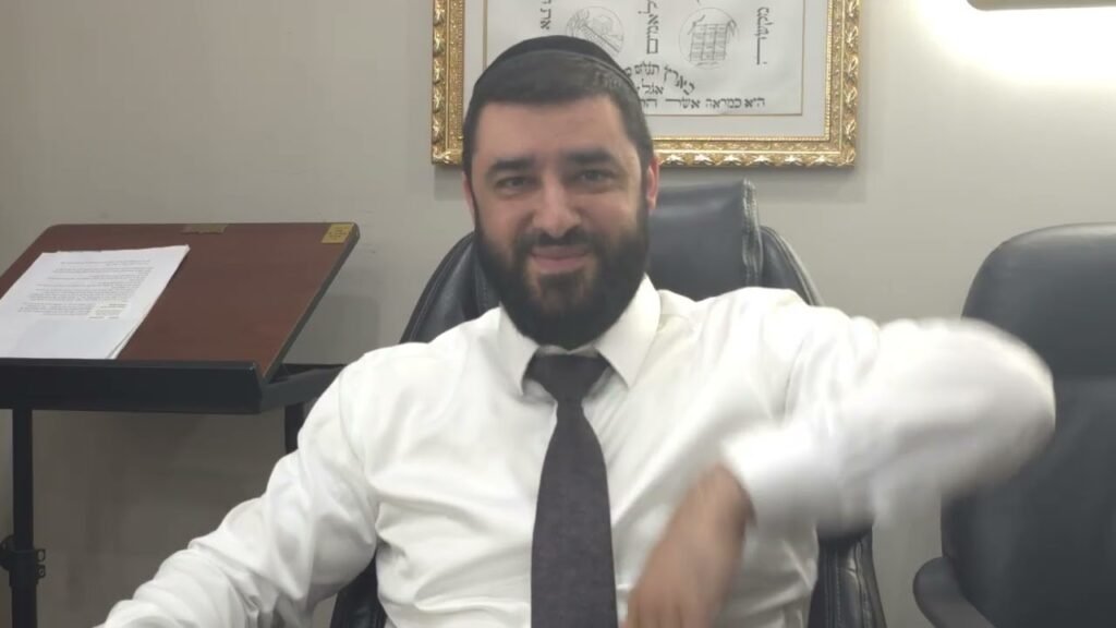 Bearded man in a white shirt and tie sits in an office chair, wearing a kippah, smiling with framed art behind him.