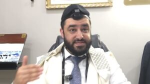 Bearded man in a kippah and tallit speaking while gesturing in an office setting.